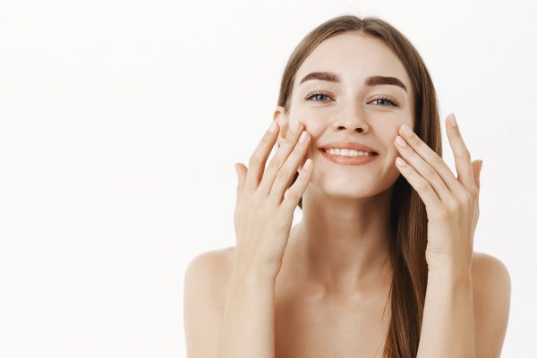Waist-up shot of charming relaxed and gentle young woman making cosmetological procedure applying facial cream on face with fingers and smiling broadly feeling perfect, taking care of skin. Beauty and skincare concept