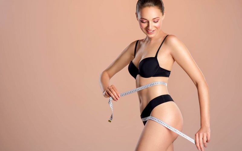Young smiling woman with tape and a beautiful, healthy body. Sexy woman measuring her perfect body. Happy girl in black underwear with an ideal body, posing with a metre. Woman measuring her waist.